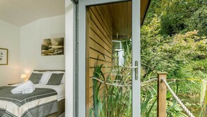 A bedroom with a bed, pillows, and a towel, a window with a view of greenery, and a wooden door. - Riverside Lodge New Forest (Godshill)