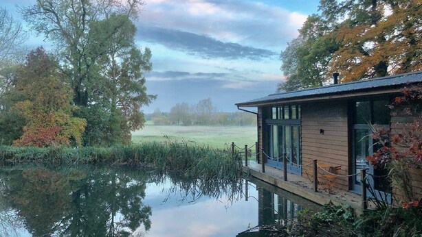 A wooden building with a glass door, surrounded by trees and a calm body of water. - Riverside Lodge New Forest (Godshill)