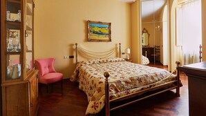 Een slaapkamer met een houten bed, een roze fauteuil, een glazen kast met decoratieve objecten, een schilderij aan de muur en een kroonluchter.