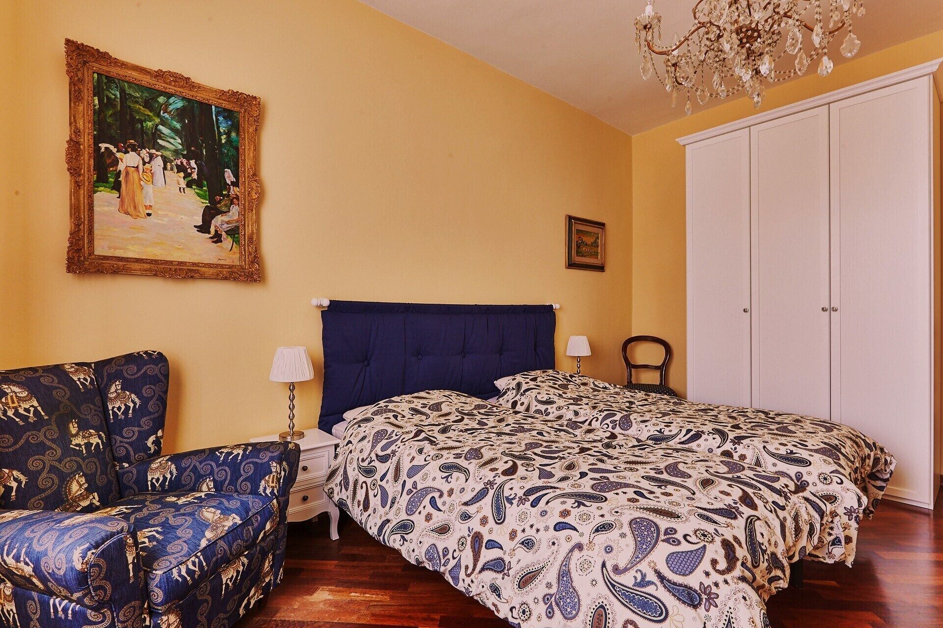 Quarto com cama, cadeira, mesa de cabeceira, abajur, pintura, guarda-roupa e lustre.