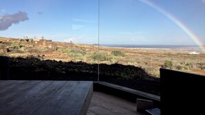Une pièce dotée d’une grande fenêtre en verre donnant sur un paysage où l’on aperçoit un arc-en-ciel, un bâtiment éloigné et un ciel dégagé.