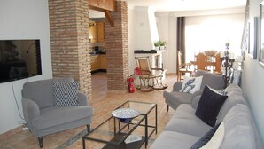 Ein Wohnzimmer mit grauem Sofa, einem Couchtisch und einem Fernseher. Im Hintergrund ist ein Kamin aus Ziegelsteinen und ein Essbereich zu sehen.