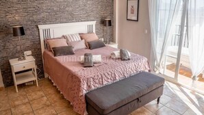 Ein Schlafzimmer mit einer Steinwand, einem Bett mit rosa Bettdecke, zwei Nachttischlampen, einem Nachttisch und einem Fenster mit durchscheinenden Vorhängen.
