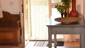 Una habitación con un banco de madera, una mesa con un florero y plantas, y un ventanal corredizo con cortina.