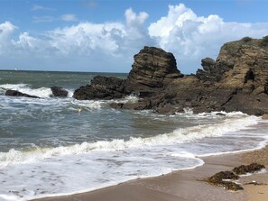 Uma praia com ondas quebrando em formações rochosas e um veleiro ao longe.