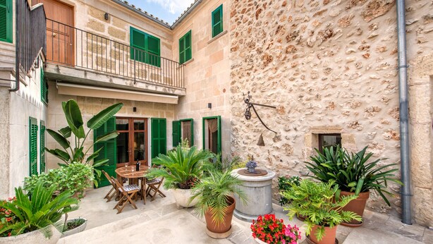 Une cour avec un mur en pierre, des volets verts, une table en bois avec des chaises et des plantes en pot.