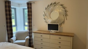 Ein Schlafzimmer mit Bett, Kommode, Sessel, Fernseher und einem Fenster mit Vorhängen.