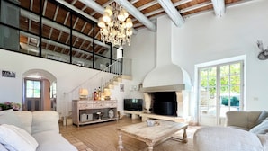 A spacious living room with a high ceiling, a fireplace, a staircase, and a large window.