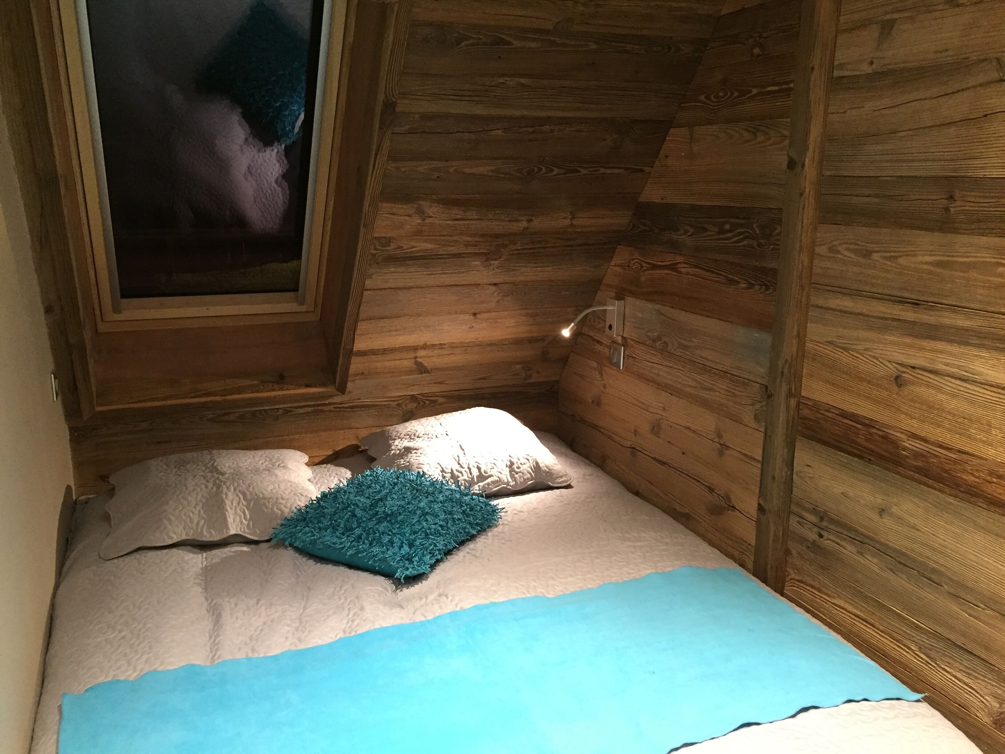 Una habitación pequeña y acogedora con paredes de madera y techo inclinado. Hay una cama con una manta azul y un almohadón, y una ventana con vistas al exterior.