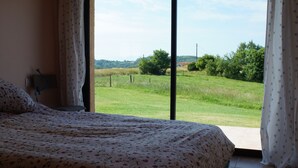 A bedroom with a large bed, a nightstand, and a view of a grassy field through a window. - Peaceful Gite + PRIVATE Pool Exclusively for you +Magnificent Panoramic Views  (Lalanne)