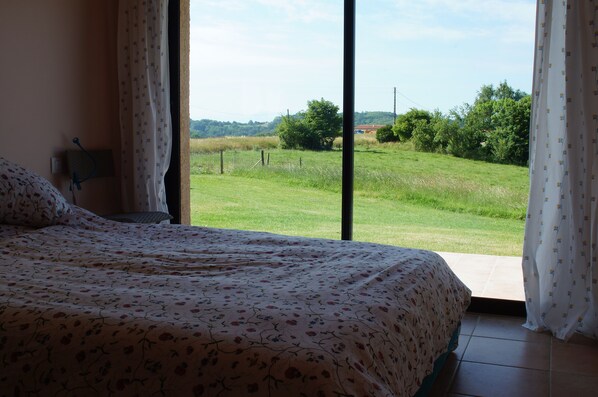 A bedroom with a large bed, a nightstand, and a view of a grassy field through a window. - Peaceful Gite + PRIVATE Pool Exclusively for you +Magnificent Panoramic Views (Lalanne)