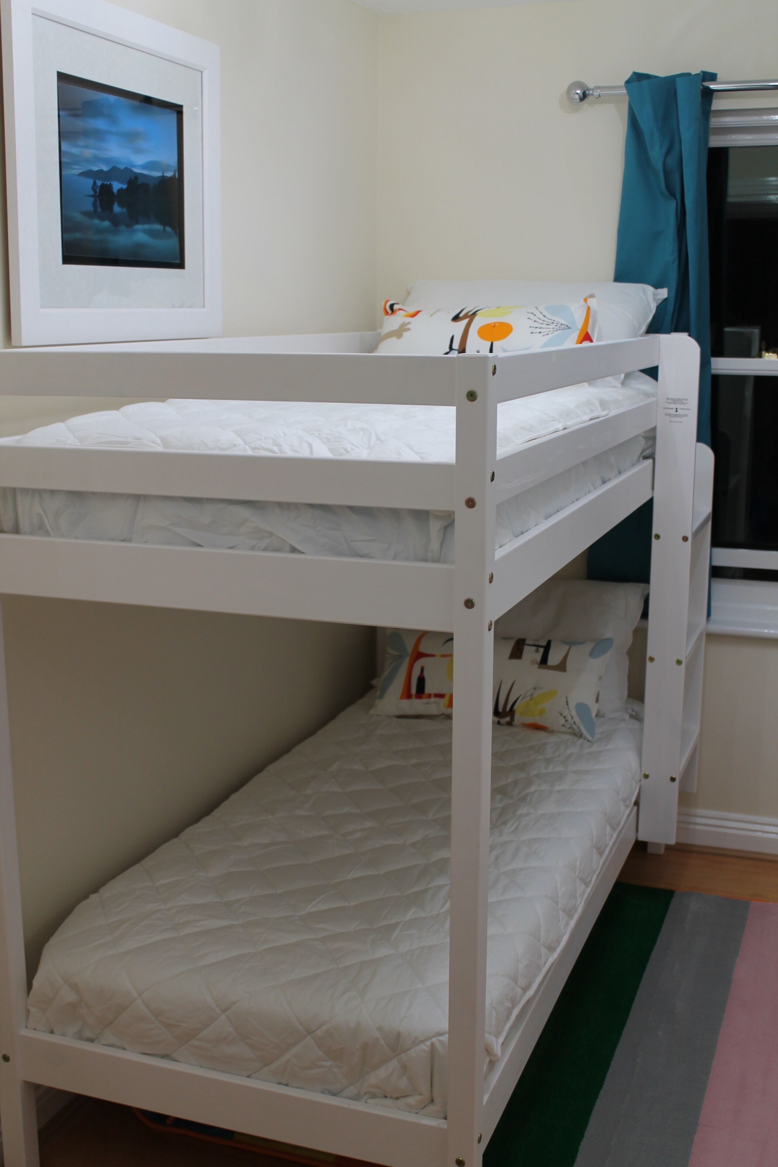 Un lit superposé avec une structure blanche, un lit inférieur doté d’un matelas matelassé et un lit supérieur avec un oreiller et une couverture.