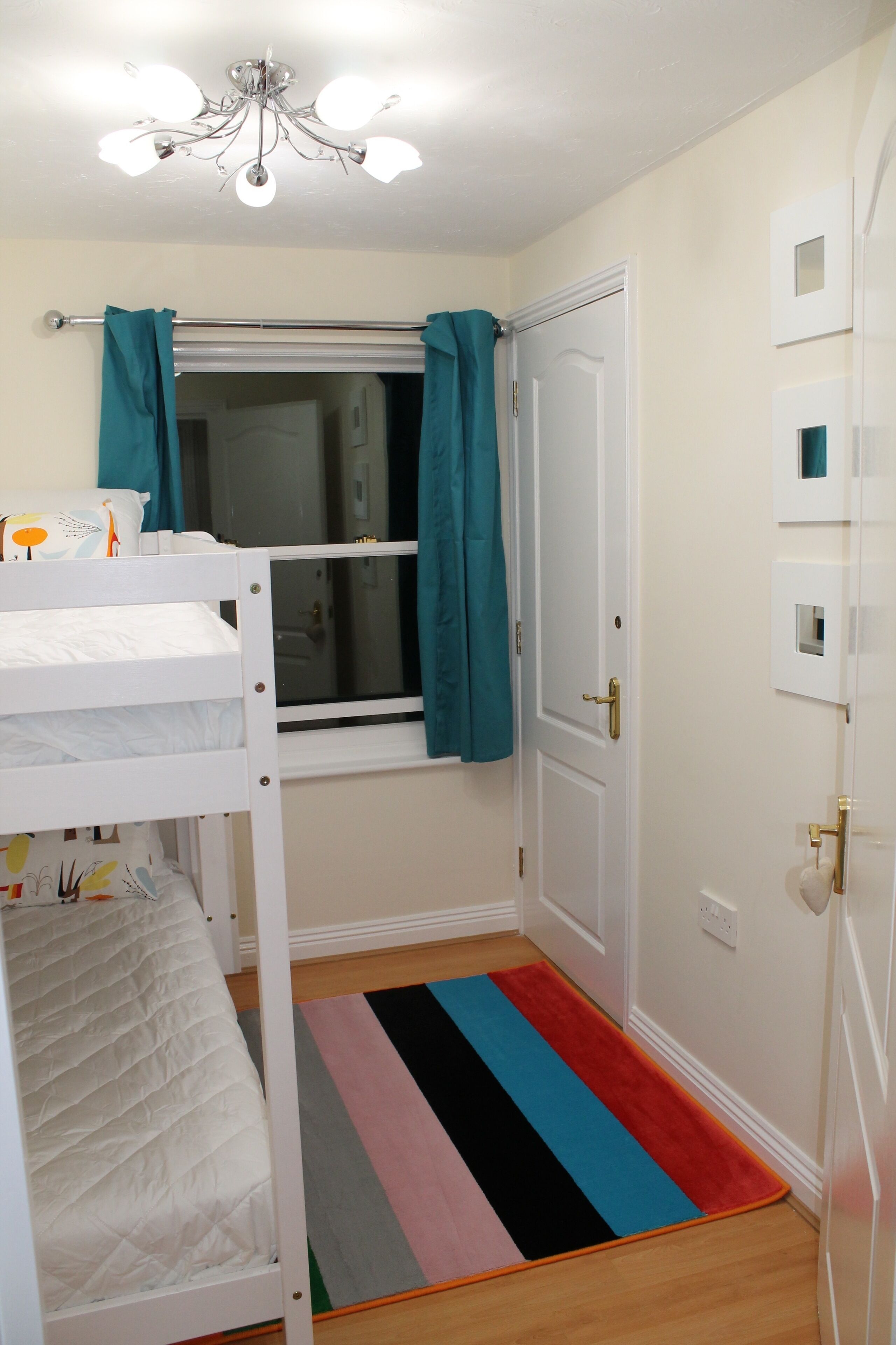 A bedroom with a bunk bed, a window with teal curtains, a door, and a colorful striped rug.