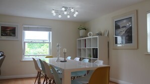 Un comedor con mesa y sillas blancas, una araña, una ventana con persianas y una estantería con objetos decorativos.