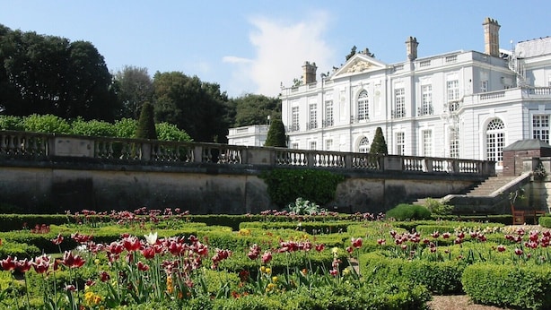 Ein formeller Garten mit verschiedenen Tulpen und ordentlich geschnittenen Hecken vor einem großen, klassizistischen Gebäude mit zahlreichen Fenstern und einer symmetrischen Fassade.