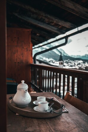 Una mesa de madera con una bandeja que sostiene una tetera y dos tazas, con el fondo de una baranda de madera y una vista nevada de la montaña.