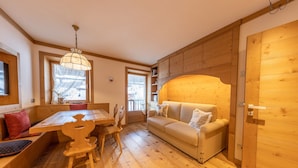 A cozy wooden interior with a dining table, benches, a sofa, and a large window.