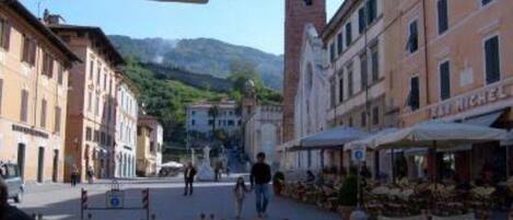 Un café al aire libre con mesas y sillas, rodeado de edificios históricos y una montaña al fondo.