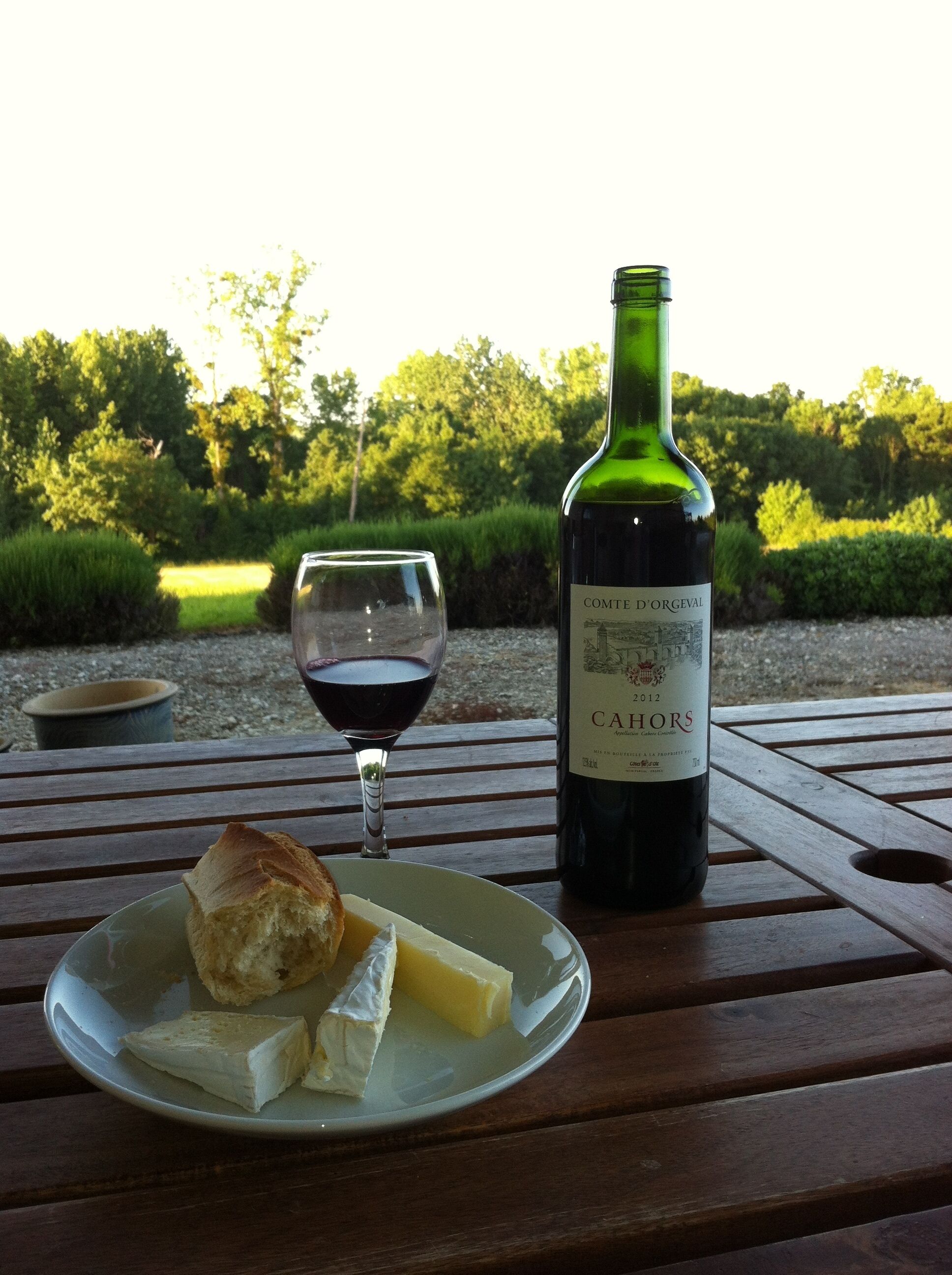 Una botella de vino tinto Comte d'Orgeval 2012 Cahors, una copa de vino tinto y un plato con queso y pan sobre una mesa de madera al aire libre.