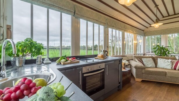 Eine Küche mit einer großen Kochinsel, einem Spülbecken und einem Backofen. Auf der Arbeitsplatte stehen Pflanzen und durch große Fenster ist ein Blick ins Freie möglich.