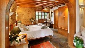 A cozy bedroom with a stone wall, wooden ceiling beams, a large bed, a bedside table with a lamp, and a window with a view of greenery. - A spacious and luxurious retreat for couples near Great Tew (Chipping Norton)