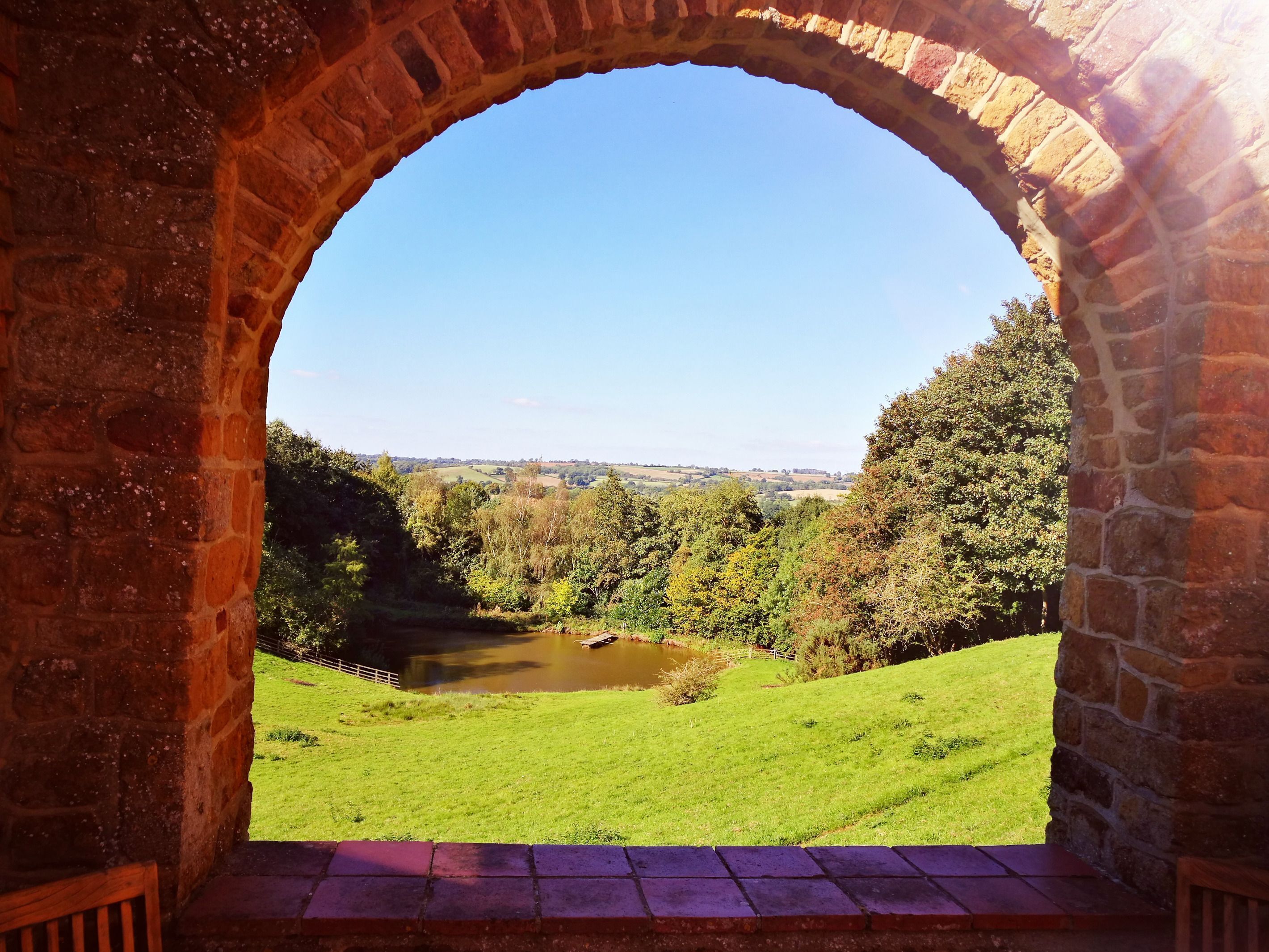 A stone archway leading to a grassy landscape with a pond and trees.