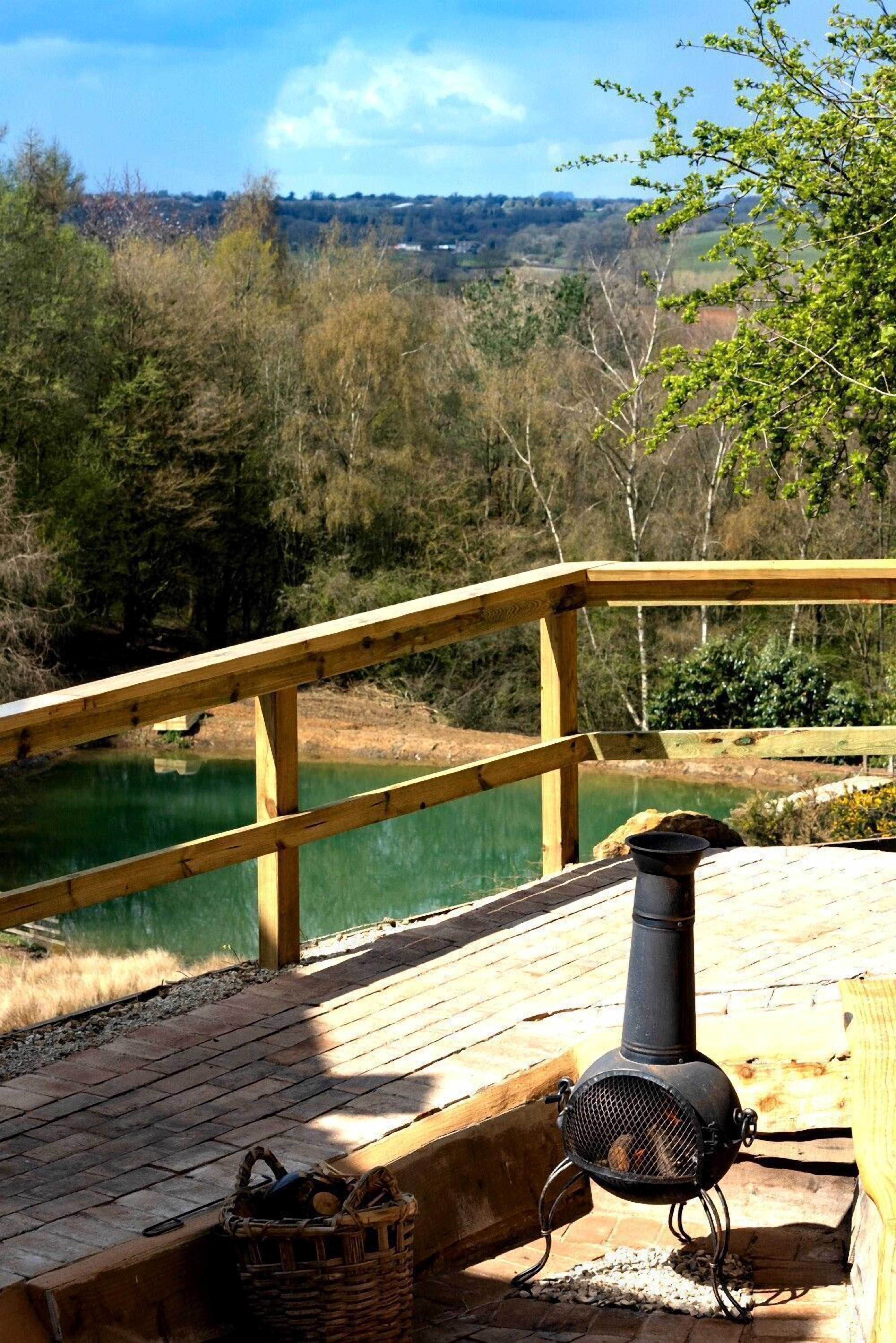 Eine Holzterrasse mit Blick auf einen See und einen Wald. Ein Schornstein und ein Holzkorb sind zu sehen.