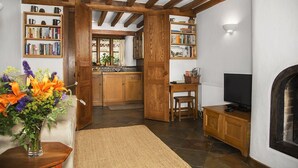 A cozy living room with a fireplace, wooden bookshelves, a television, and a kitchen area visible through an open door. - A spacious and luxurious retreat for couples near Great Tew (Chipping Norton)