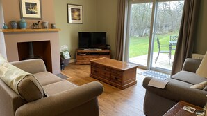 Ein Wohnzimmer mit zwei Sofas, einem Holztisch, einem Kamin, einem Fernseher und Blick auf einen Garten durch eine Schiebetür.