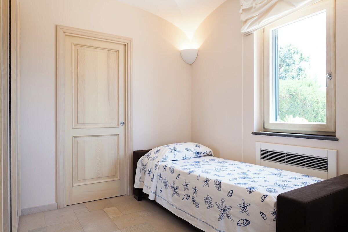 A small, single-bed room with a window, a wall-mounted light, and a door.