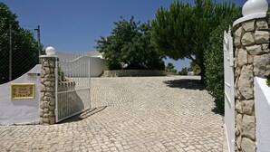 Un camí empedrat que condueix a una entrada amb tanca, reforçada per un mur de pedra i rematada amb un pinacol esfèric.
