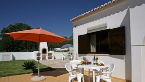 Un pati exterior amb una taula i caires blancs, un parasol vermell i un edifici blanc amb una finestra i un persianes.