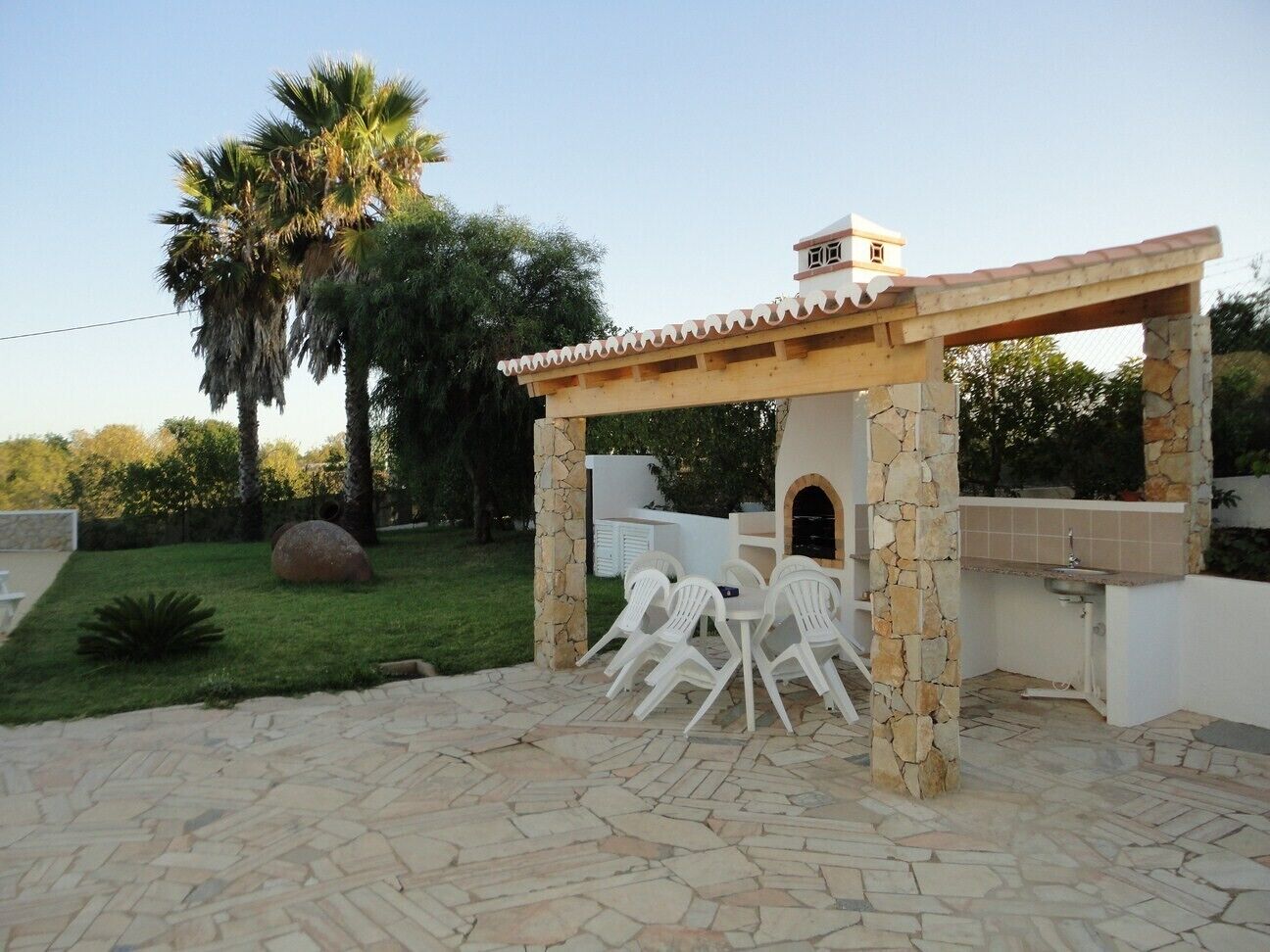 An outdoor patio with stone flooring, a pergola structure, a stone-built barbecue area, and a seating arrangement with white plastic chairs.