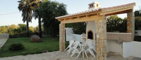 An outdoor patio with stone flooring, a pergola structure, a stone-built barbecue area, and a seating arrangement with white plastic chairs.