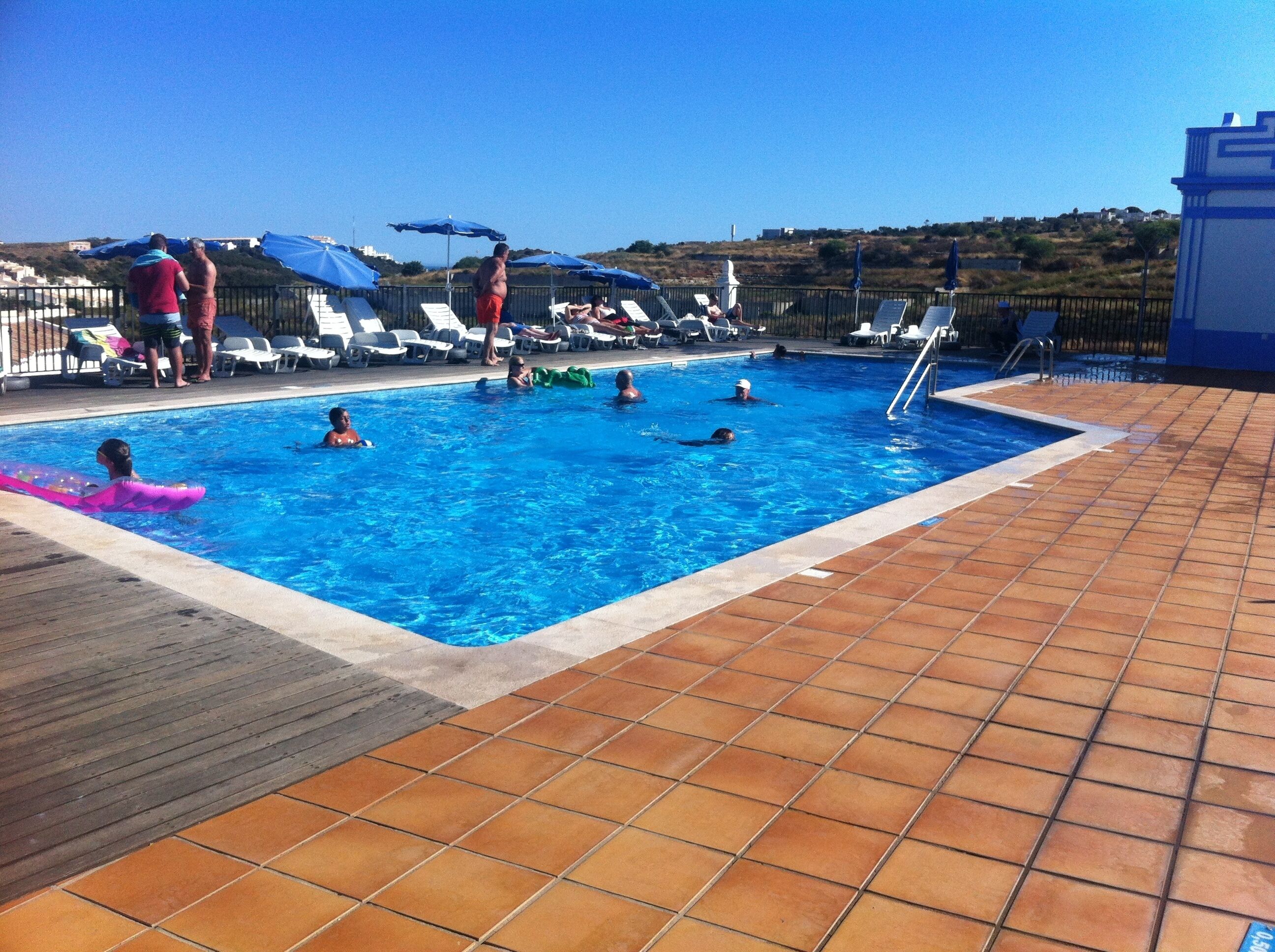 A swimming pool with people in it, surrounded by lounge chairs and umbrellas.