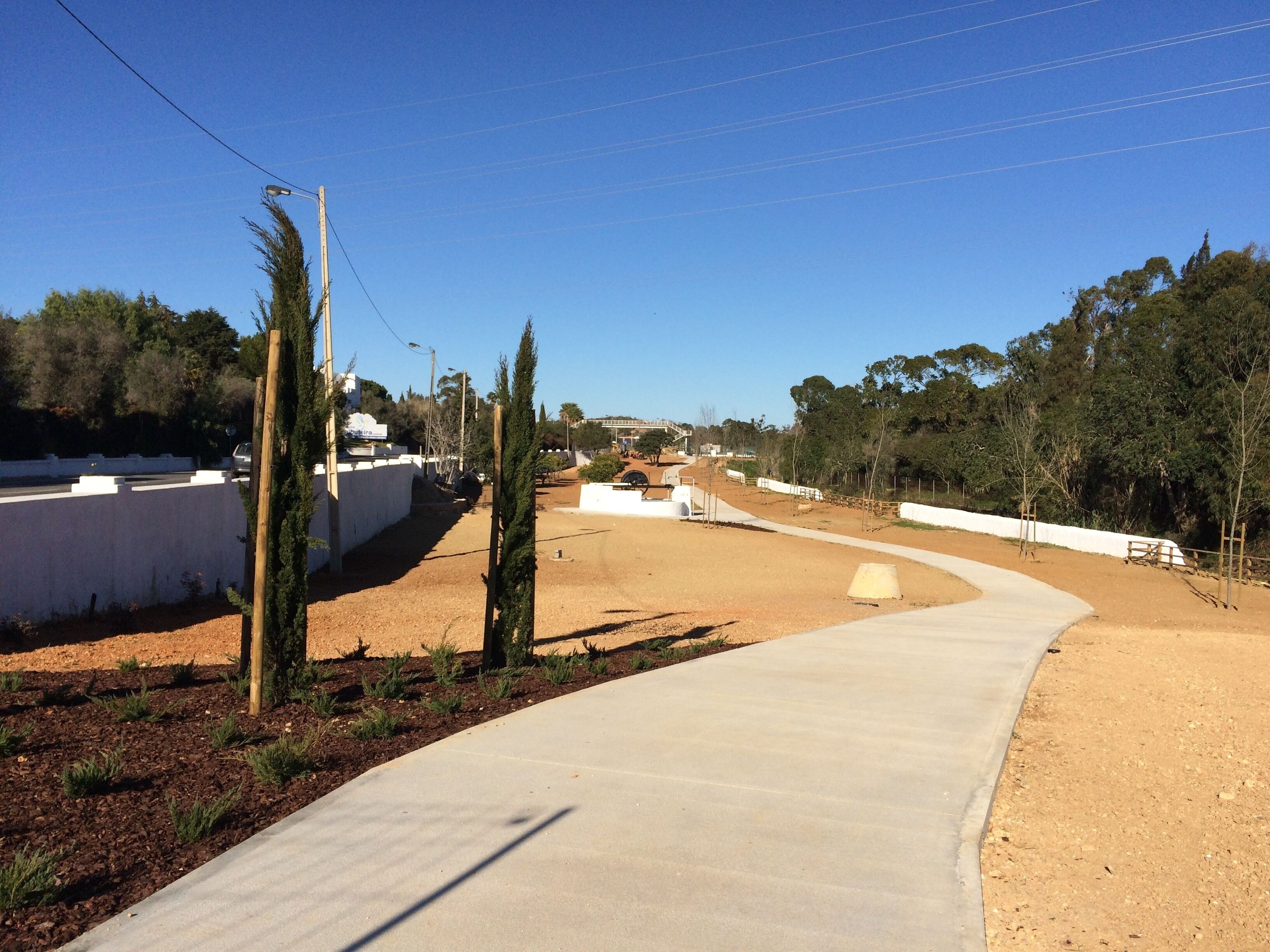 A paved pathway curving through a landscaped area with a white wall on one side and trees on the other.