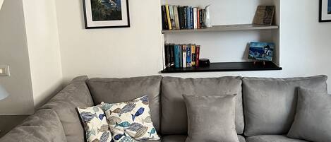 A modern living room with a grey sofa, a bookshelf with books and decorative items, a framed picture on the wall, and a lamp.