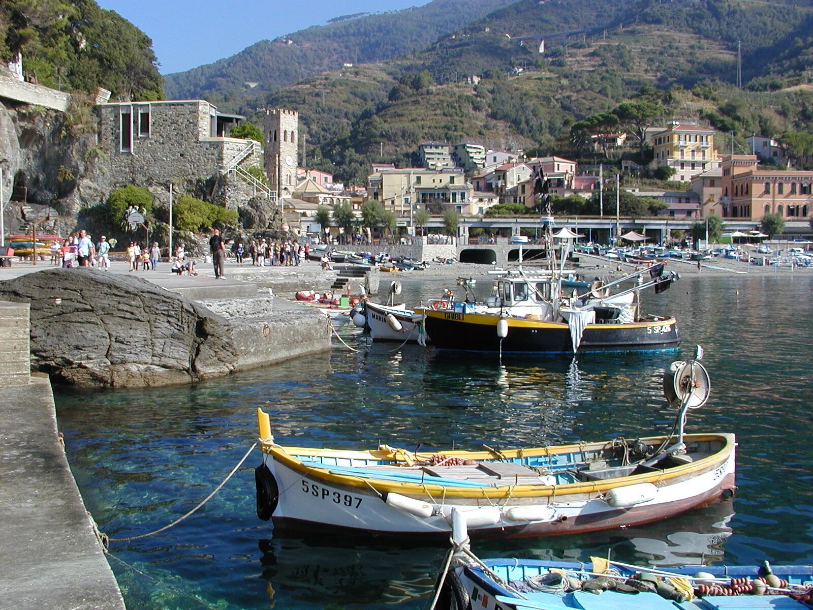 A coastal town with boats docked at a pier, stone buildings, and a mountainous backdrop.