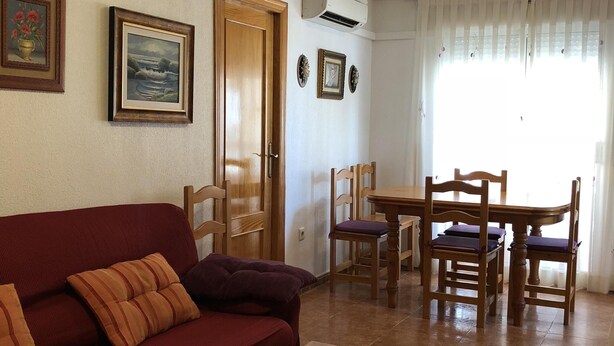 Una sala de estar con un sofá rojo, una mesa de madera y sillas. Hay una mesa de comedor con sillas, una puerta y cuadros en la pared.