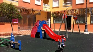 Un parque infantil con tobogán y columpios frente a un edificio con balcones y un cartel que dice 'DIRECT'.