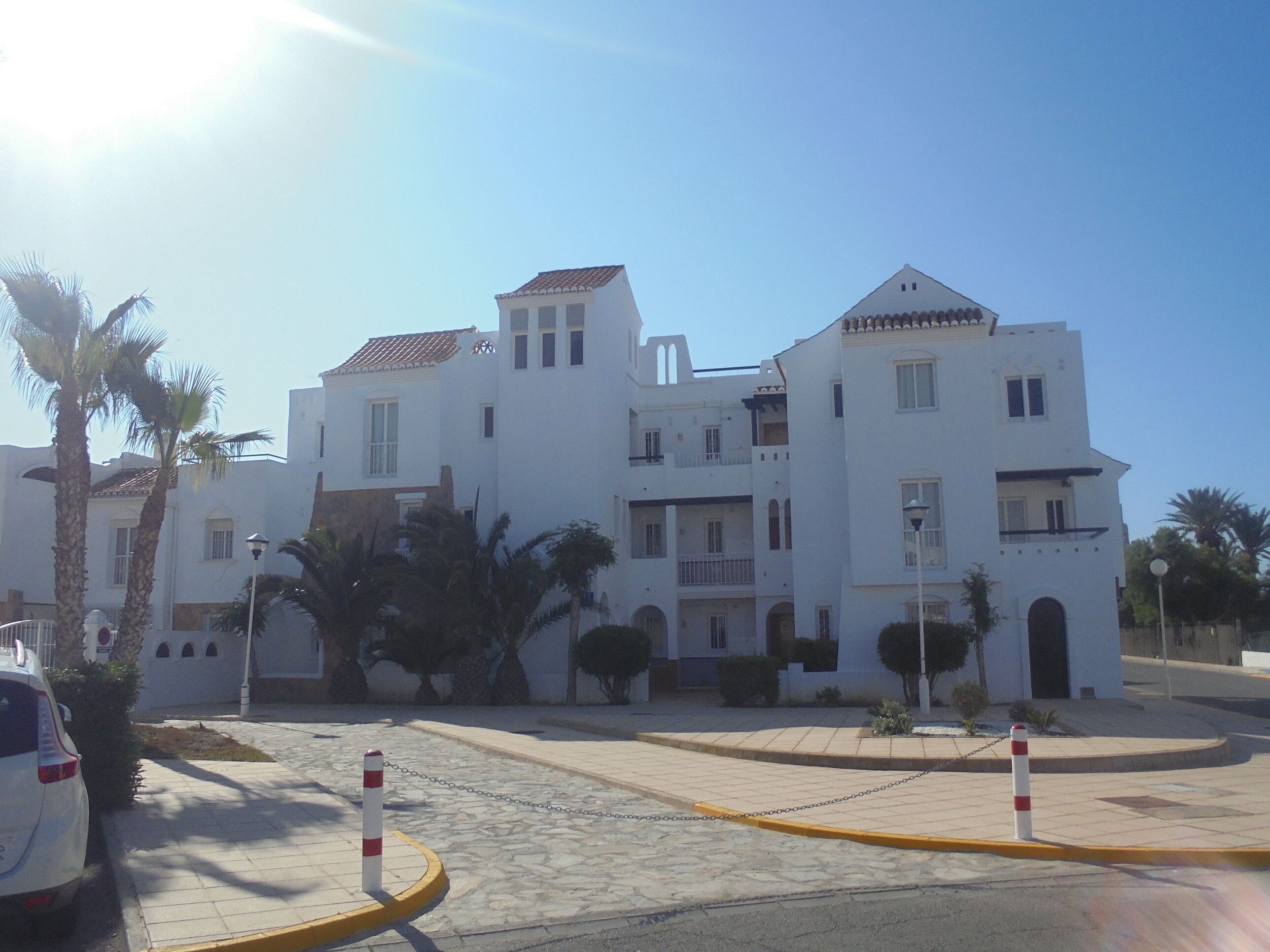 A white building with multiple balconies and a tiled roof, surrounded by palm trees and a paved area with bollards.