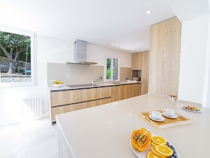 A modern kitchen with a central island, wooden cabinets, and a view of greenery outside. - Villa Alcanada Sunrise. Your villa with private pool and sea views in Alcanada. (Alcúdia)