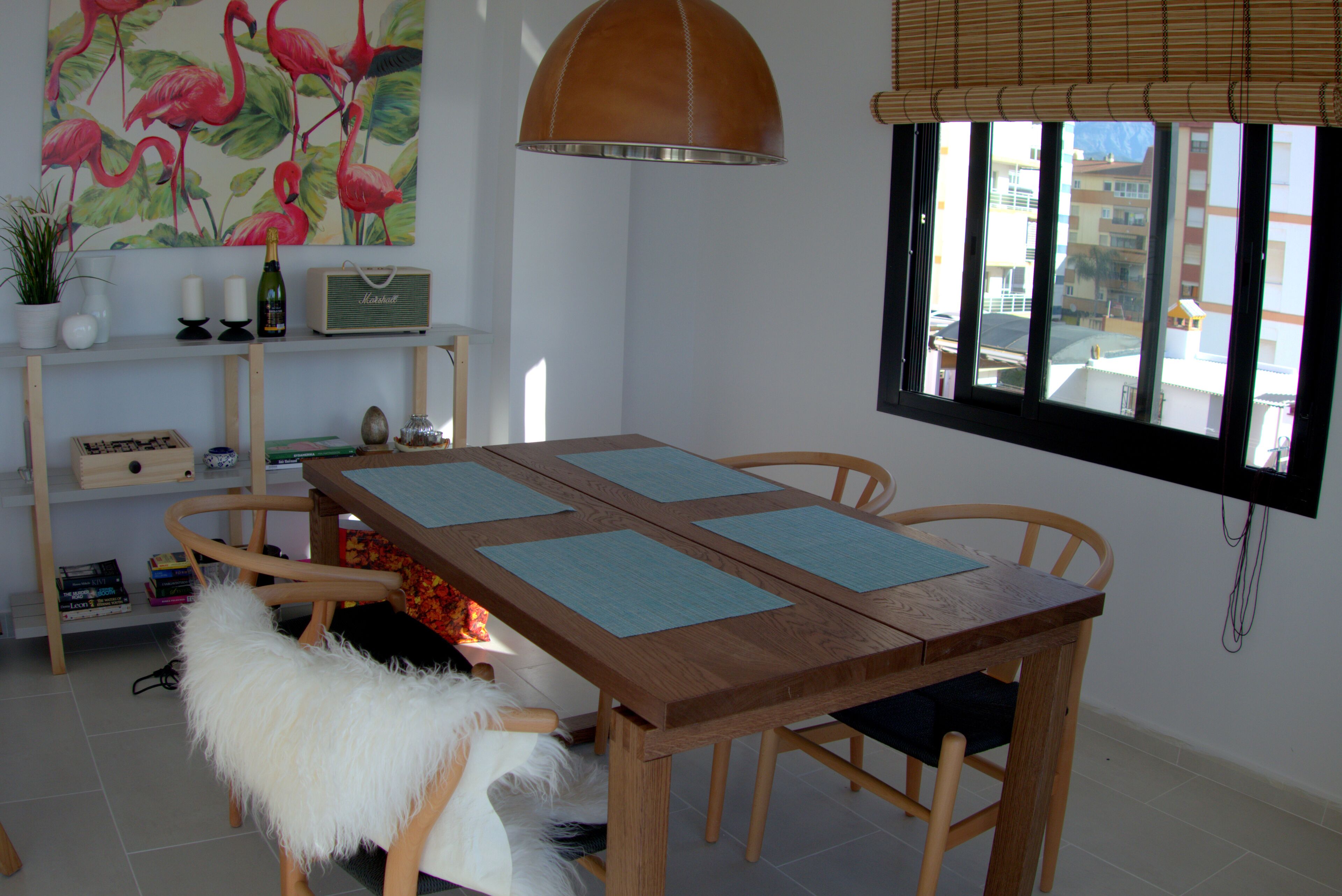 Un coin repas avec une table en bois, quatre chaises, un fauteuil blanc moelleux, une bibliothèque remplie de livres et d’objets décoratifs, un tableau représentant des flamants roses et une fenêtre donnant sur des immeubles.