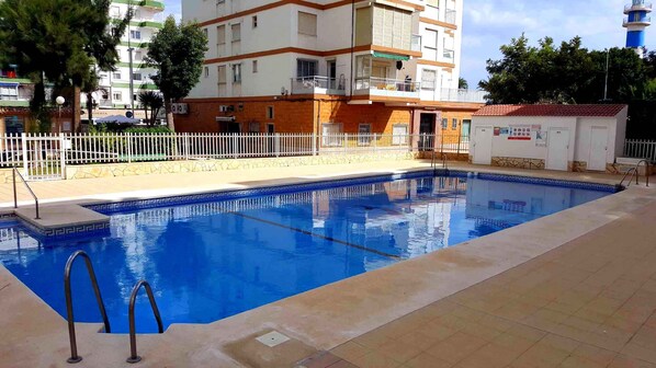Uma piscina com cerca branca, um pequeno edifício com um letreiro e uma construção alta ao fundo.