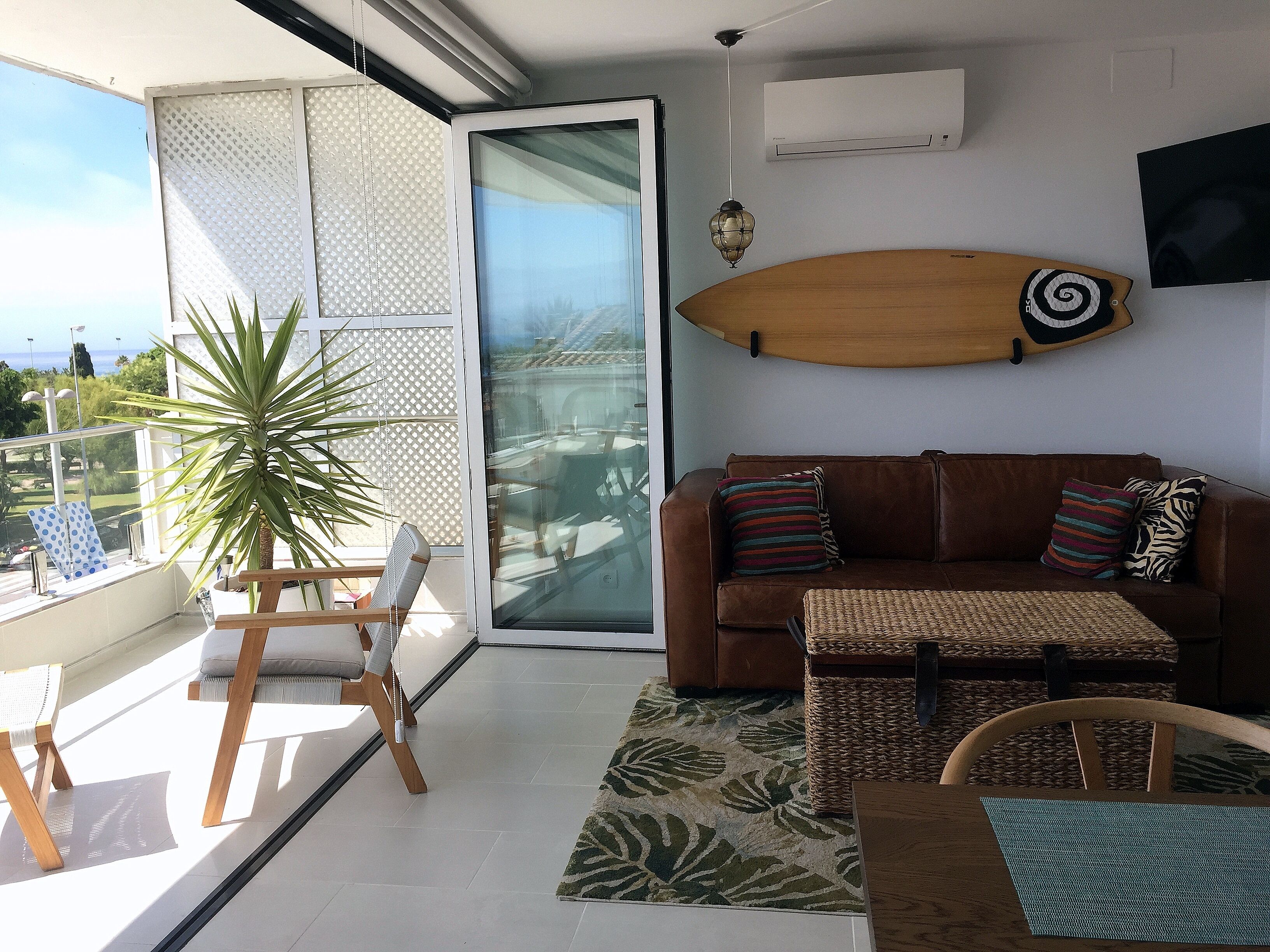 Un soggiorno moderno con un divano in pelle marrone, una porta vetrata che dà su un balcone, un'asse da surf appesa al muro e una pianta in vaso.