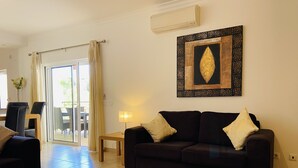 A living room with a dark-colored sofa, a wooden coffee table with a shell-patterned plate, a lamp, and a wall-mounted air conditioner. - Beautiful 2 Bed Apartment with Swimming Pool in the Historic Town of Lagos (Lagos)