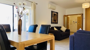 A modern living room with a wooden dining table, black chairs, a blue sofa, and a dark-colored couch. There is a vase with flowers on the table, and a painting on the wall. - Beautiful 2 Bed Apartment with Swimming Pool in the Historic Town of Lagos (Lagos)