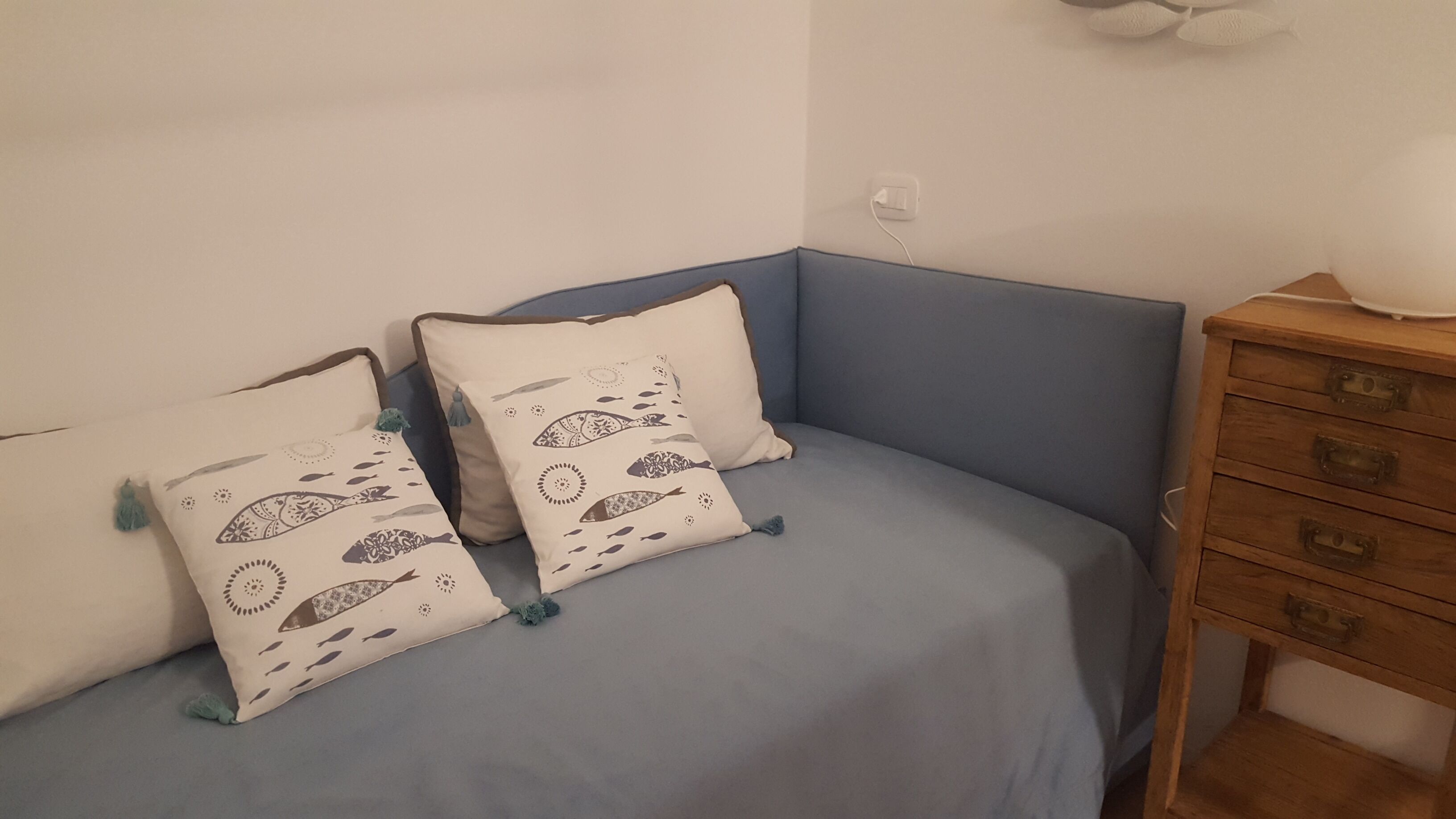 Une petite pièce chaleureuse, avec un canapé bleu et deux coussins à motifs. À droite, on aperçoit une commode en bois avec des tiroirs.