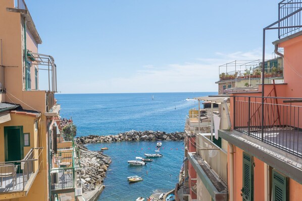 A narrow coastal street with buildings on either side, a rocky breakwater, and boats in the water.