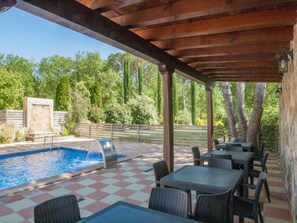 Un espace extérieur avec piscine, doté d’un sol à carreaux, entouré de tables et de chaises, et surplombé d’une pergola en bois.